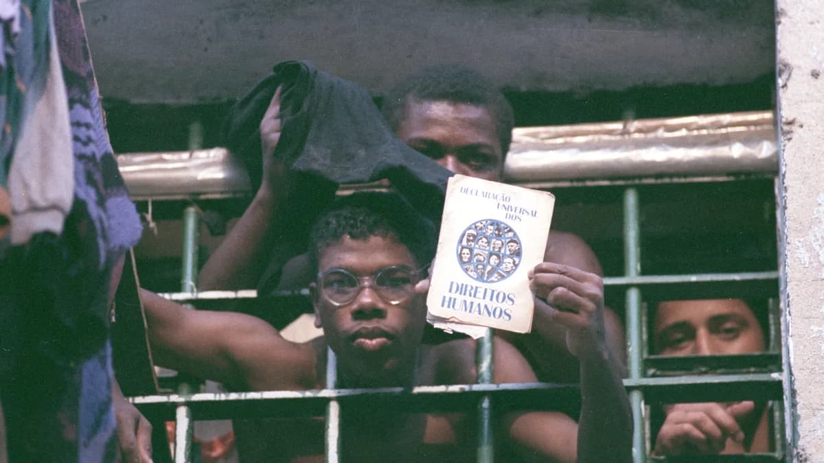 Texto alternativo: Homens presos aparecem atrás das grades de uma cela. Em primeiro plano, um deles segura um exemplar da Declaração Universal dos Direitos Humanos diante das grades, enquanto outros observam ao fundo. A fotografia registra o interior de uma unidade prisional e destaca a relação entre encarceramento e direitos humanos.