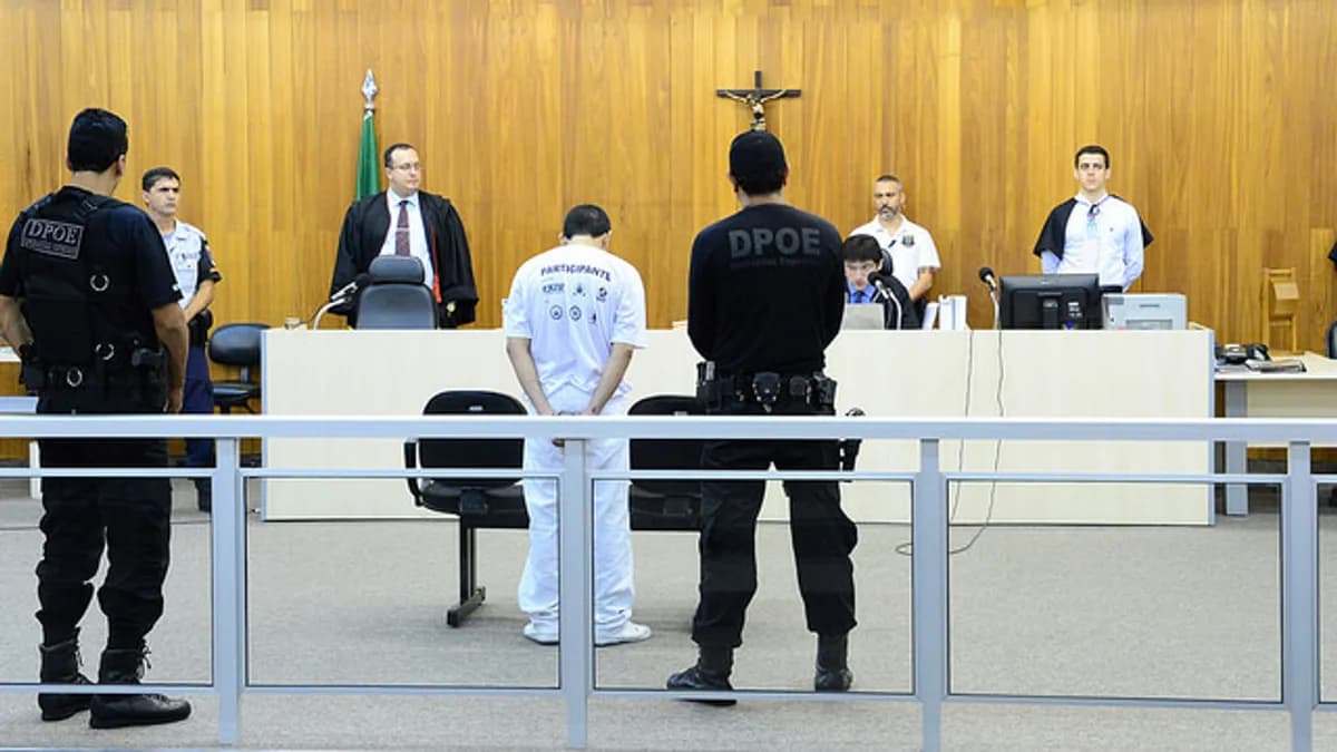 ala de audiência do Tribunal do Júri, com um réu em pé diante do plenário, acompanhado por agentes do Departamento de Polícia Especializada (DPOE). Ao fundo, integrantes do sistema de Justiça ocupam a bancada sob um crucifixo.