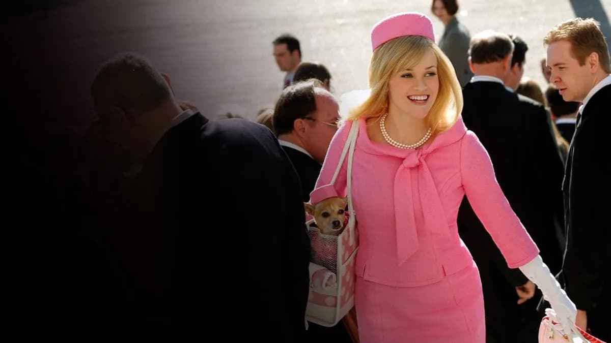 Cena de Legalmente Loira, com Elle Woods usando um conjunto rosa vibrante, chapéu combinando, colar de pérolas e luvas brancas, enquanto carrega uma bolsa com seu cachorro. Ela caminha sorrindo entre várias pessoas vestidas de preto e cinza. Legalmente Loira (2001) | Prime Video.