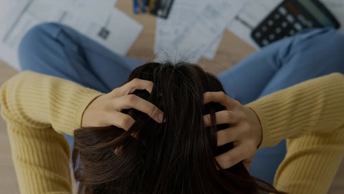 Pessoa vista de cima, sentada no chão com as mãos na cabeça diante de documentos fiscais espalhados, cartões de crédito e uma calculadora, em cena que representa dívidas tributárias